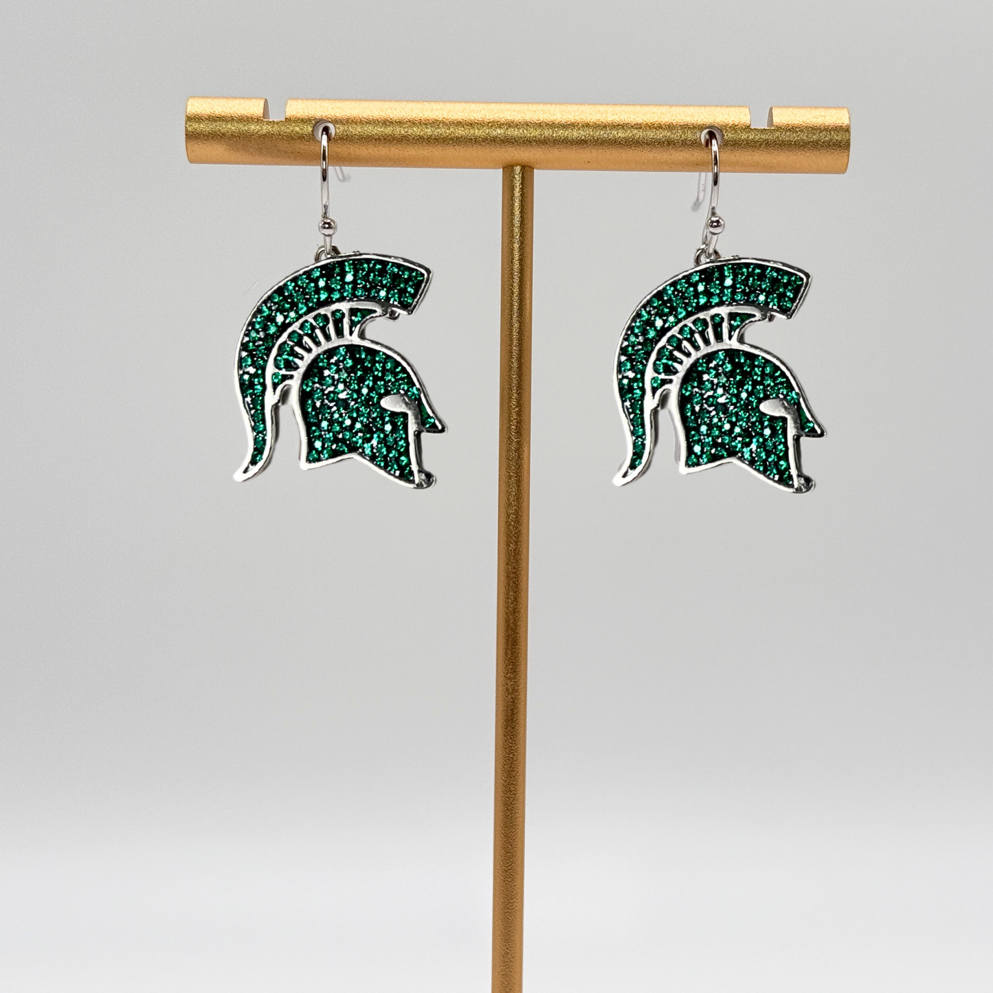 A pair of dangle earrings shaped as a Spartan Helmet logo. The logo is comprised of small green crystals with a rhodium plated brass outline.