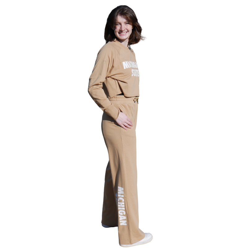 A woman wearing a tan cropped crewneck sweatshirt with matching sweatpants. On the sweatshirt reads "Michigan State" in white. On the side of the right leg of the sweatpants also reads Michigan State in white. 