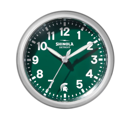 Close up of the Shinola desk clock with a green face, wooden base, and silver rim. The numbers are white and there is a white Spartan Helmet logo in place of the number 6.
