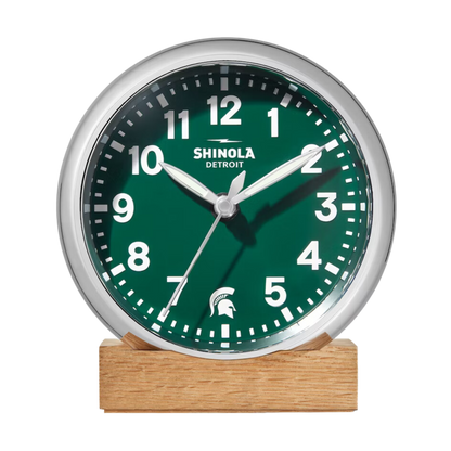 Front of the Shinola desk clock with a green face, wooden base, and silver rim. The numbers are white and there is a white Spartan Helmet logo in place of the number 6.