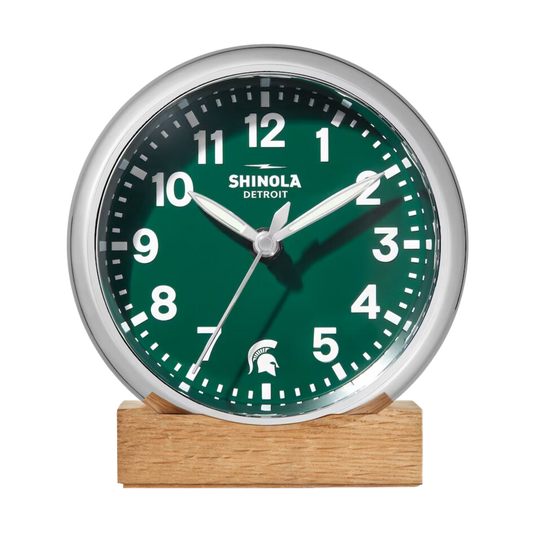 Front of the Shinola desk clock with a green face, wooden base, and silver rim. The numbers are white and there is a white Spartan Helmet logo in place of the number 6.