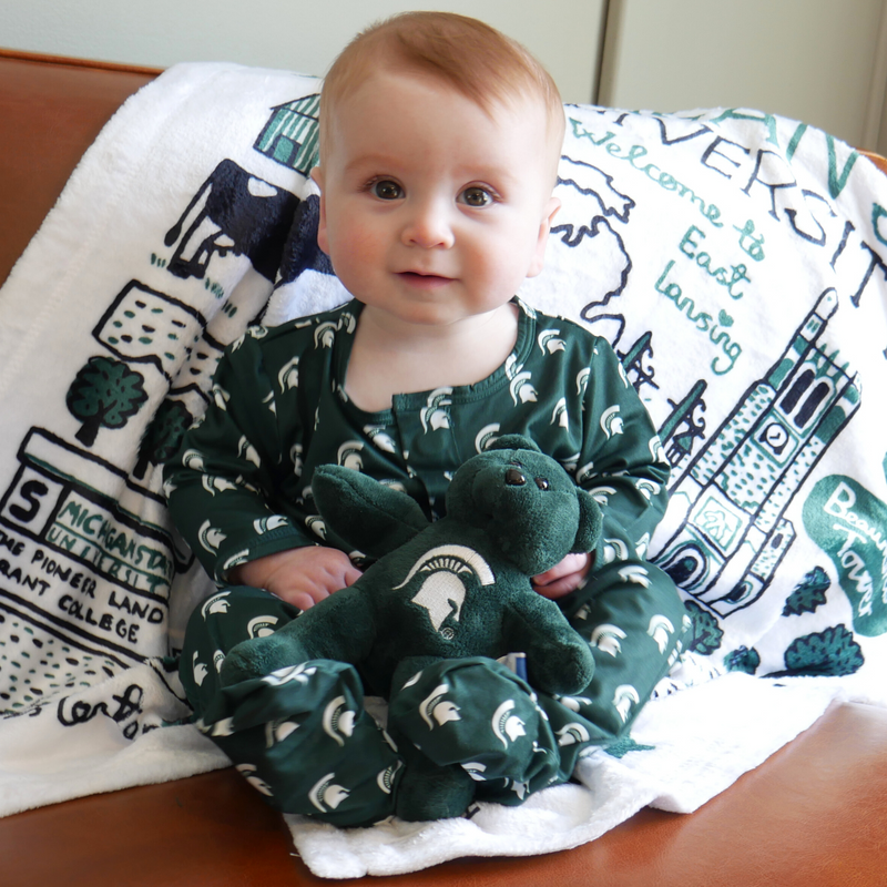 A toddler wearing a green footie onesie with small Michigan State Spartan helmet logos scattered throughout the onesie in white, while holding a green Michigan State teddy bear. 