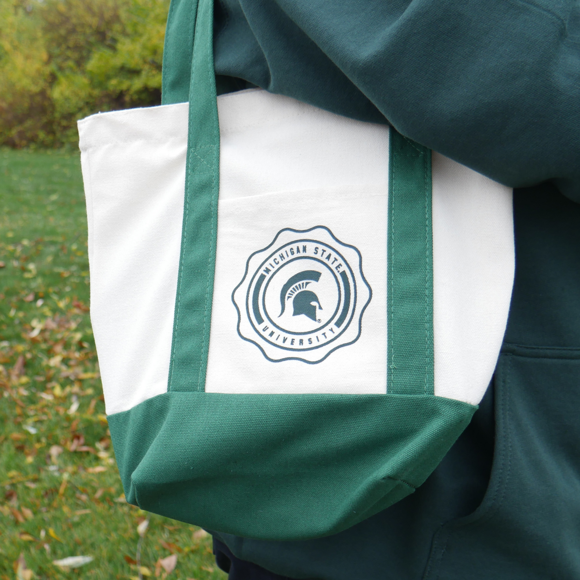 Close up of a woman carrying a cream colored canvas tote bag with dark green handles and a dark green bottom. In the middle of the bag is a circular emblem with the words "Michigan State University" around the border. In the middle of the emblem is a green Spartan Helmet logo. 