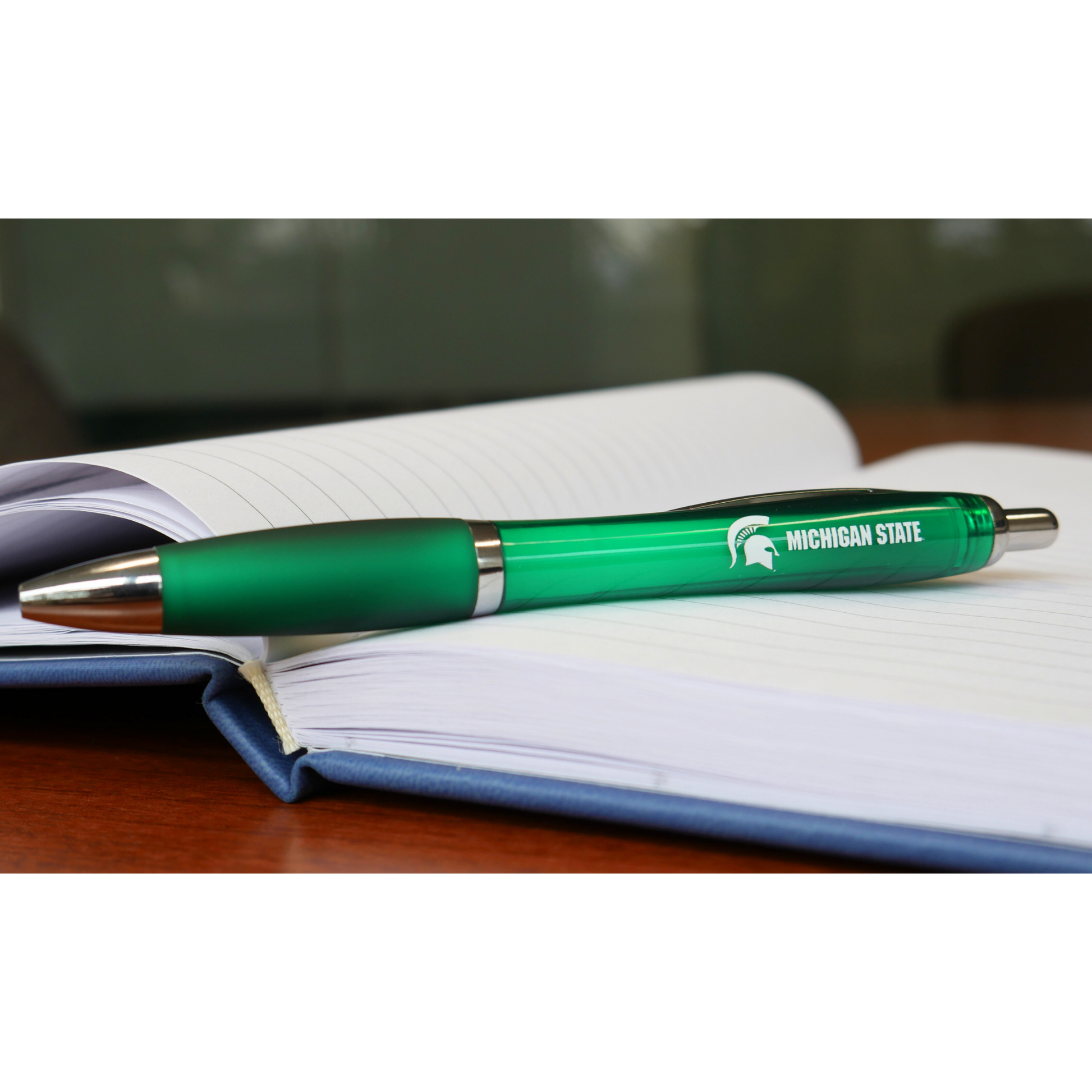 A green ballpoint pen. On the side of the pen, in white all caps text are the words "Michigan State." To the left of the text is a white Spartan Helmet logo. The pen is sitting on top of an open business notebook. 