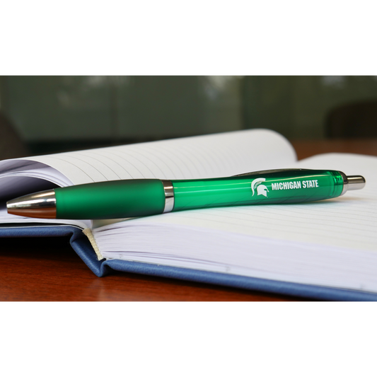 A green ballpoint pen. On the side of the pen, in white all caps text are the words "Michigan State." To the left of the text is a white Spartan Helmet logo. The pen is sitting on top of an open business notebook. 
