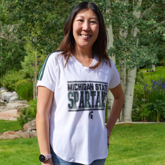 A woman standing in a backyard wearing a white, curved hem V-neck T-shirt with dual green stripes on the sleeves. The torso of the shirt reads Michigan State Spartans in two shades of green, while underneath the text is a green Spartan Helmet logo. 