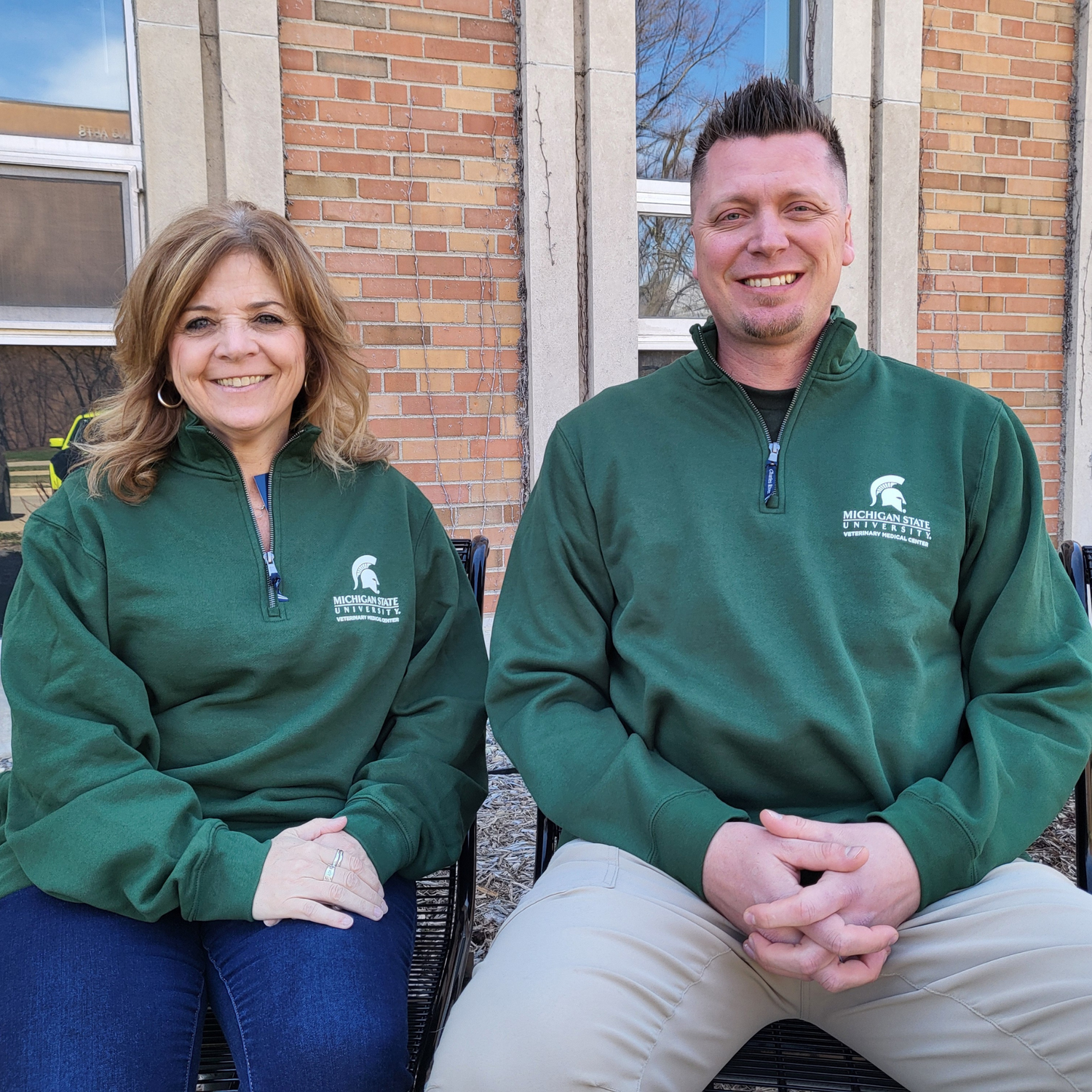 A man and a woman wearing a dark green, quarter-zip sweatshirt. The upper left chest is embroidered with a white Spartan helmet logo and Michigan State University Veterinary Medical Center logo.