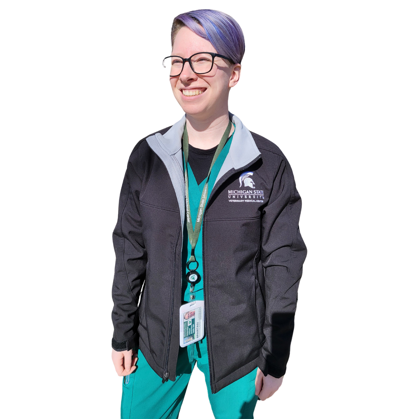 A woman in teal scrubs wearing a black full-zip jacket with a contrast light gray inside lining. The upper left chest is embroidered with a Spartan Helmet logo that is centered over the Michigan State University wordmark logo with block letters reading “Veterinary Medical Center” beneath.