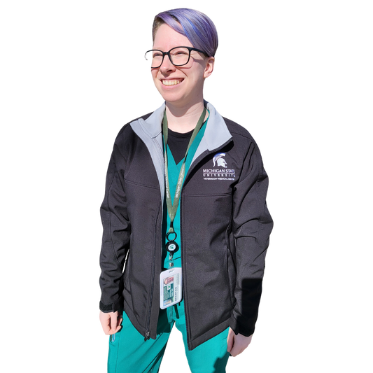 A woman in teal scrubs wearing a black full-zip jacket with a contrast light gray inside lining. The upper left chest is embroidered with a Spartan helmet logo that is centered over the Michigan State University wordmark logo with block letters reading “Veterinary Medical Center” beneath.