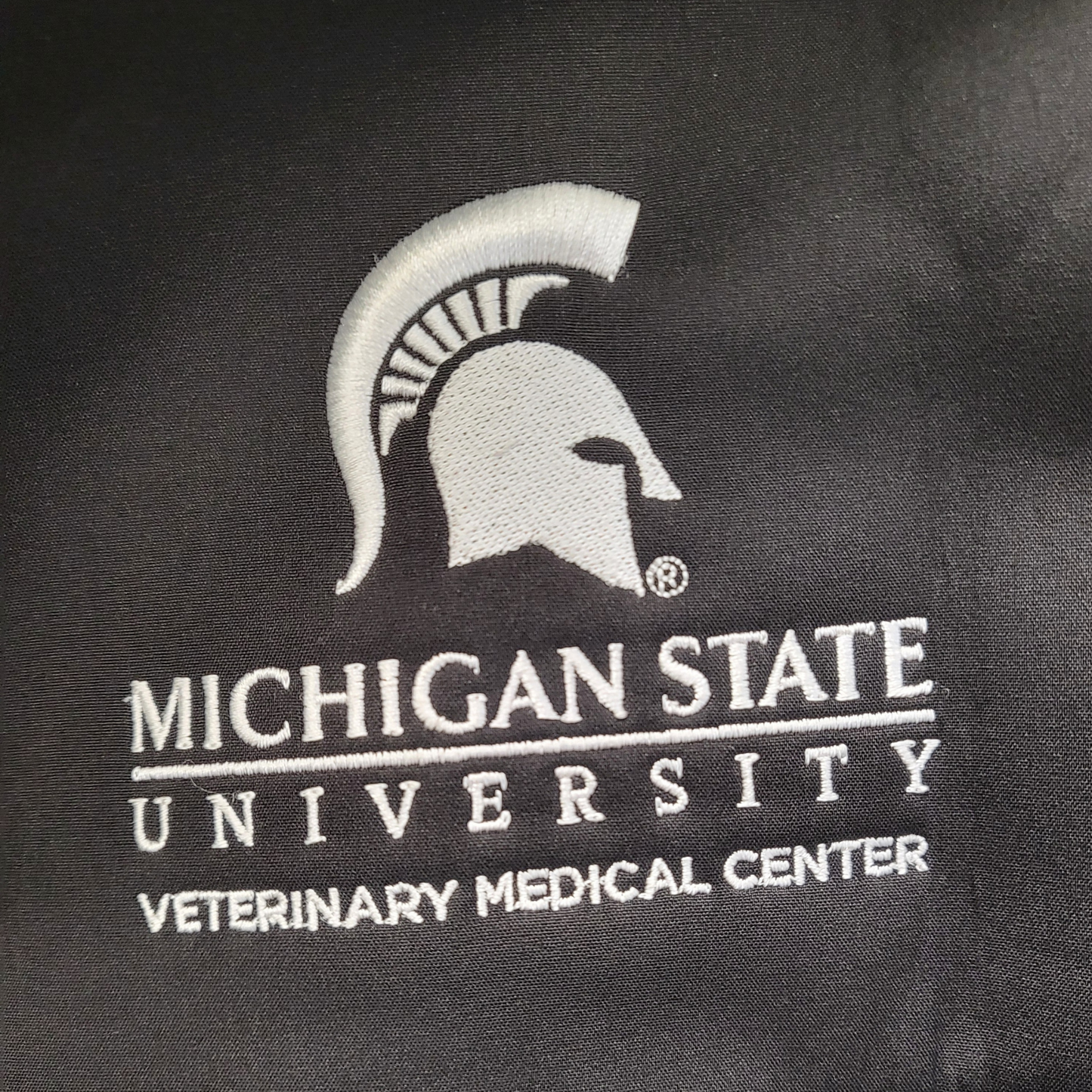 Close up of the left chest that features a white embroidered Spartan Helmet logo that is centered over the Michigan State University wordmark logo with block letters reading “Veterinary Medical Center” beneath.