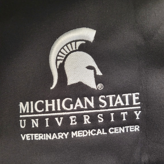 Close up of the left chest, featuring an embroidered white Spartan Helmet logo that is centered over the Michigan State University wordmark logo with block letters reading “Veterinary Medical Center” beneath. 