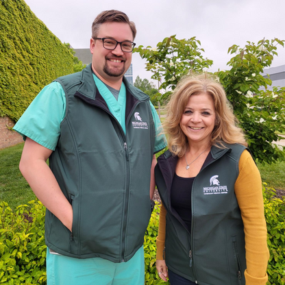 Two people wearing a green zippered vest with a white embroidered College of Osteopathic Medicine logo on the left upper chest. 
