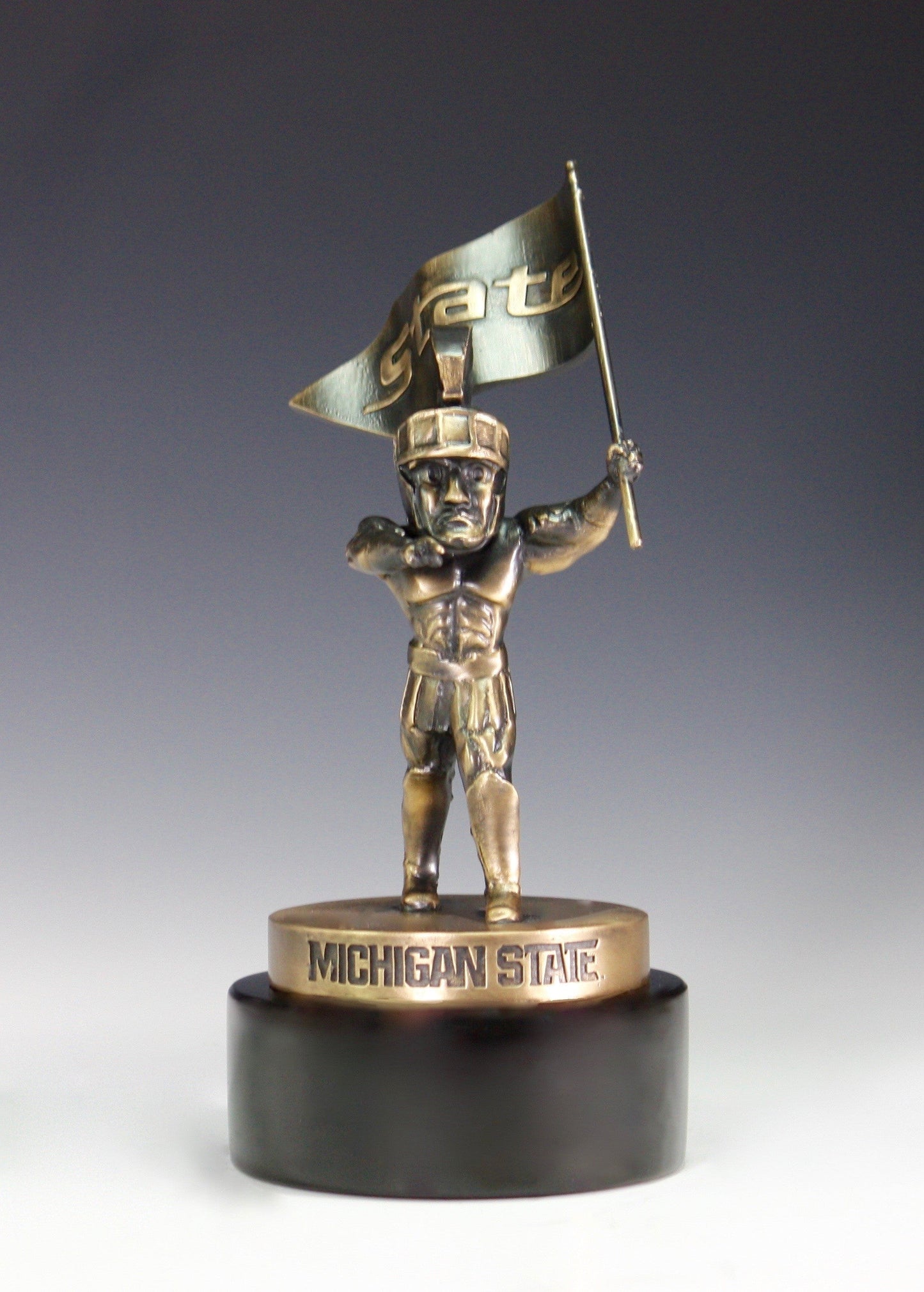 A bronze Sparty statue holding a pennant flag with the word "State" on a black base.