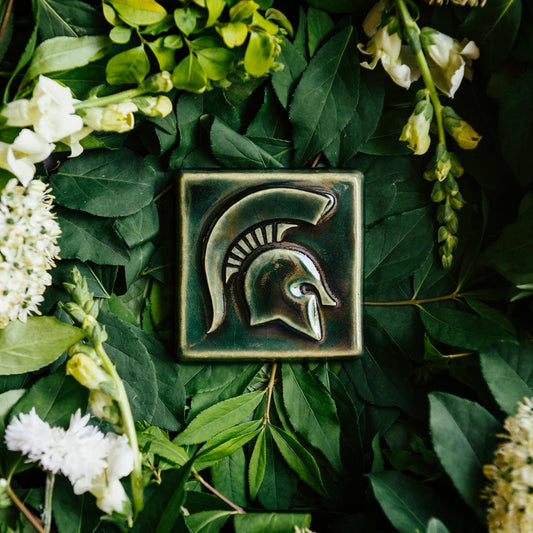 Green ceramic tile with a Spartan Helmet design, surrounded by green leaves and white flowers