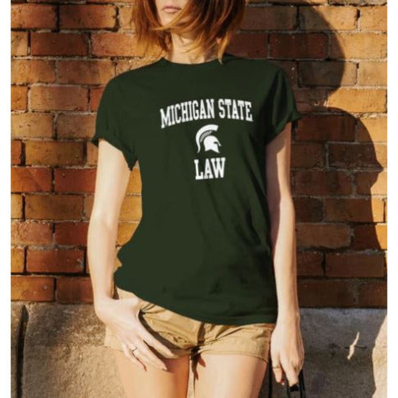 Dark green t-shirt with 'MICHIGAN STATE' in white arched text above a Spartan helmet logo, and the word 'LAW' in white below the logo. The shirt is worn by a person standing against a brick wall, paired with beige shorts.