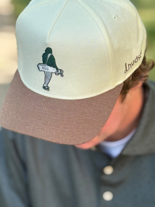 Person showing the left side of a two-tone beige and brown cap with an icon of a person holding a golf bag printed on the front and the brown d.hudson logo on the left side of the hat.  
