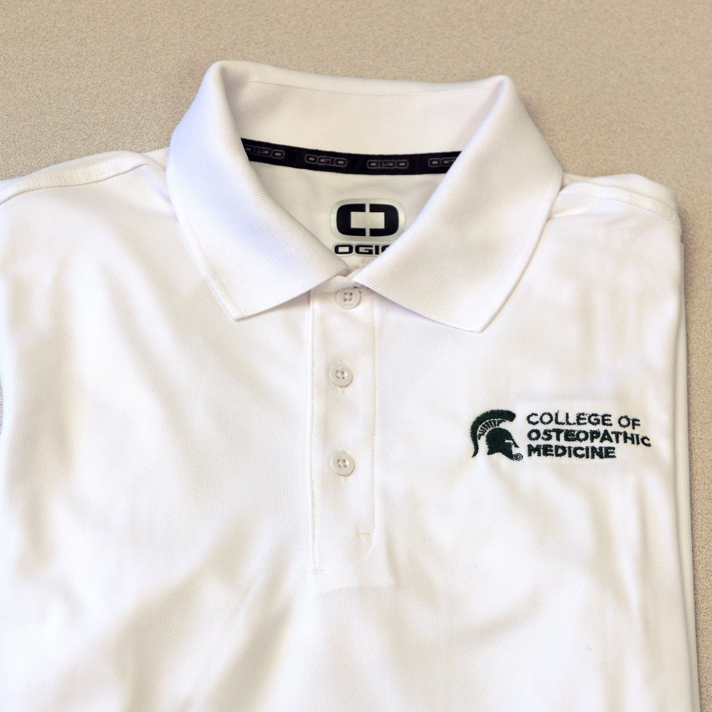 Front of white three white button polo with a green embroidered College of Osteopathic Medicine logo on the left side upper chest. 
