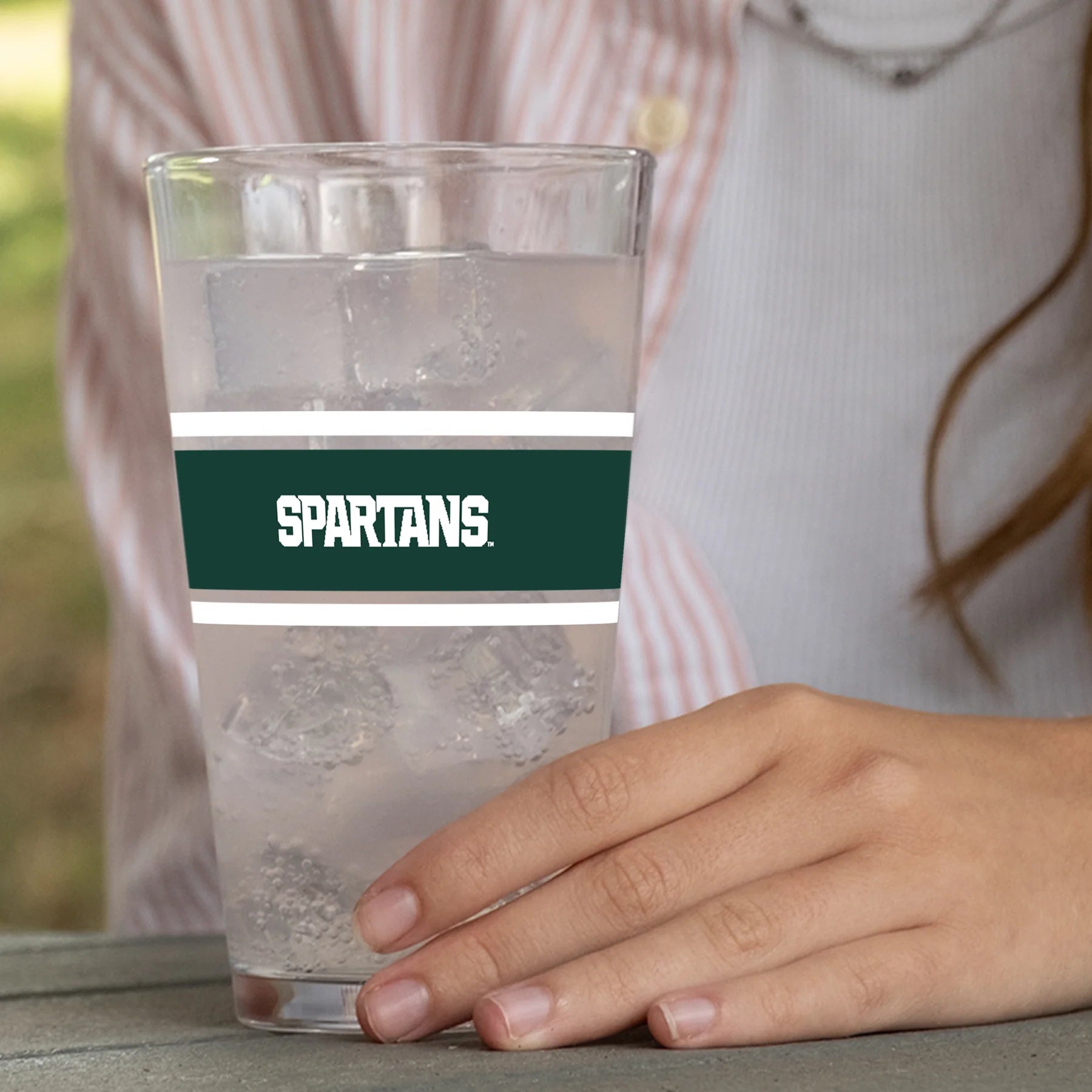 Pint glass cup with two white stripes and a thicker green stripe in the middle. On top of the green stripe is a Spartans wordmark in white. The cup is pictured with a beverage inside and a person holding it. 