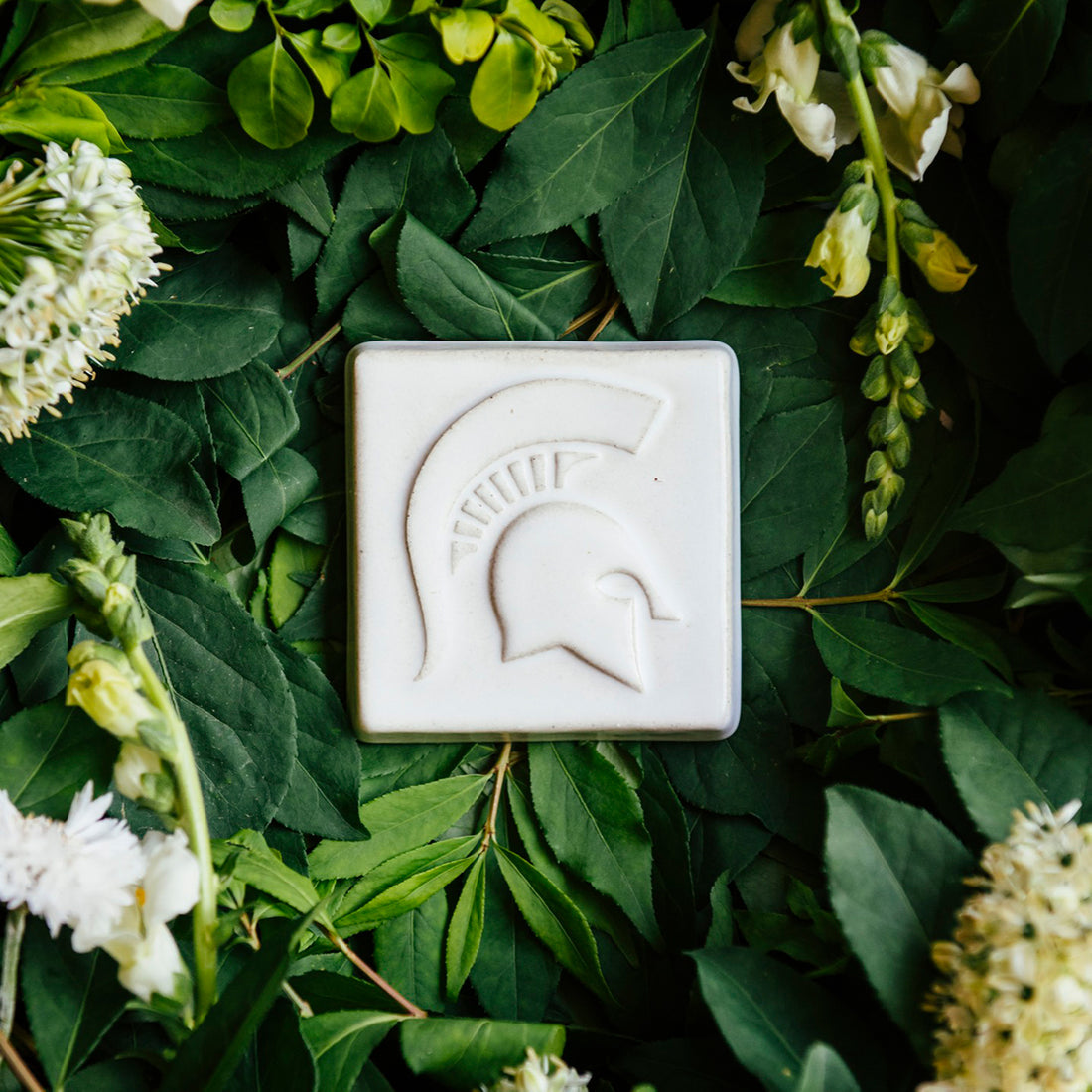 White square tile with a Spartan Helmet design, shown on a bed of green leaves and white flowers