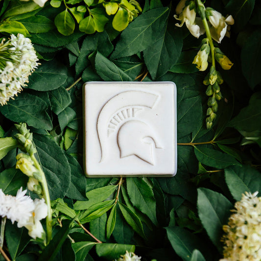 White square tile with a Spartan Helmet design, shown on a bed of green leaves and white flowers